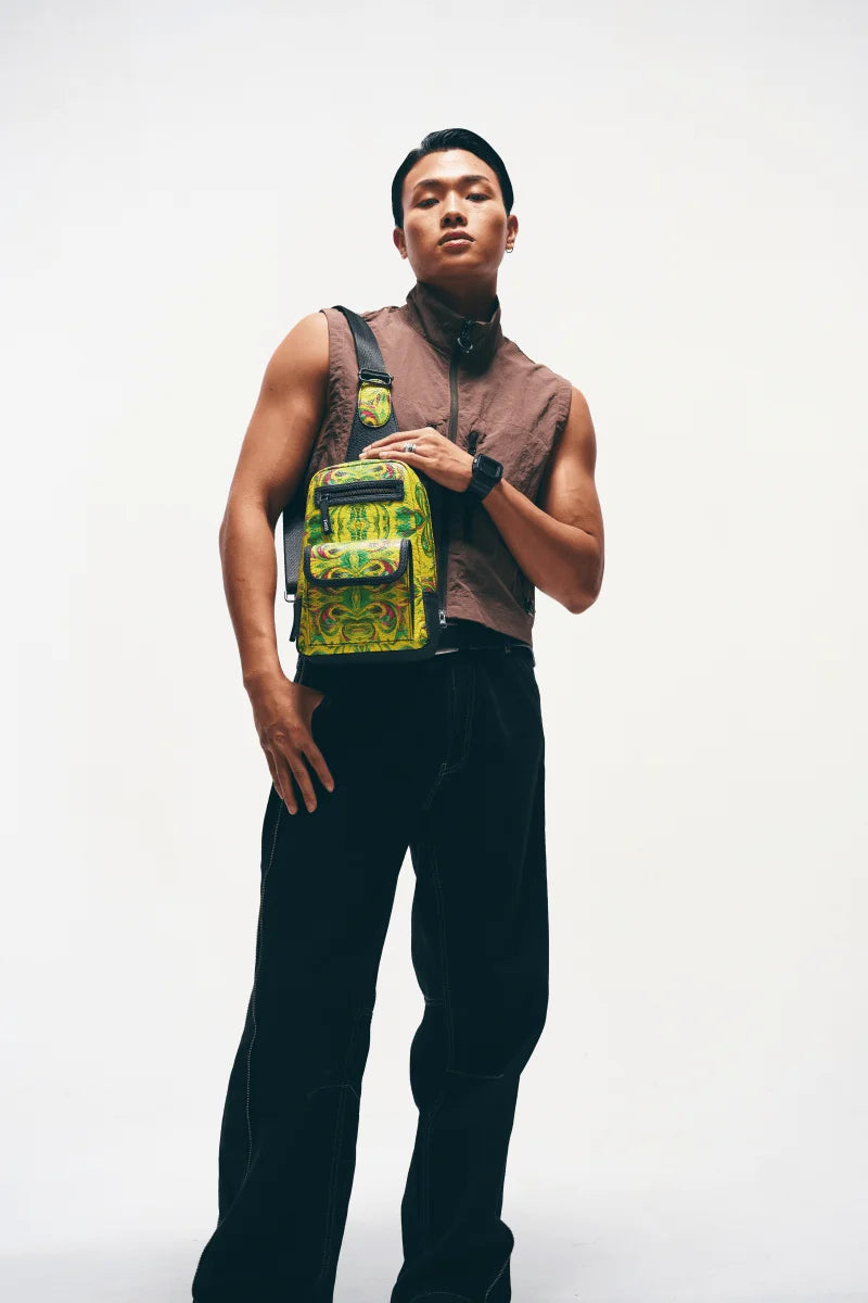 A Man wearing a brown sleeveless shirt and black pants with a Kosmic Immersion helios sling bag over his shoulder on a white background.