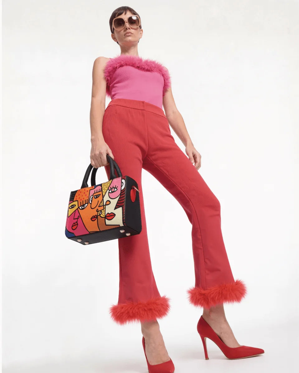 Woman in pink top and red pants holding a Mini FETTI Faces handbag on a white background
